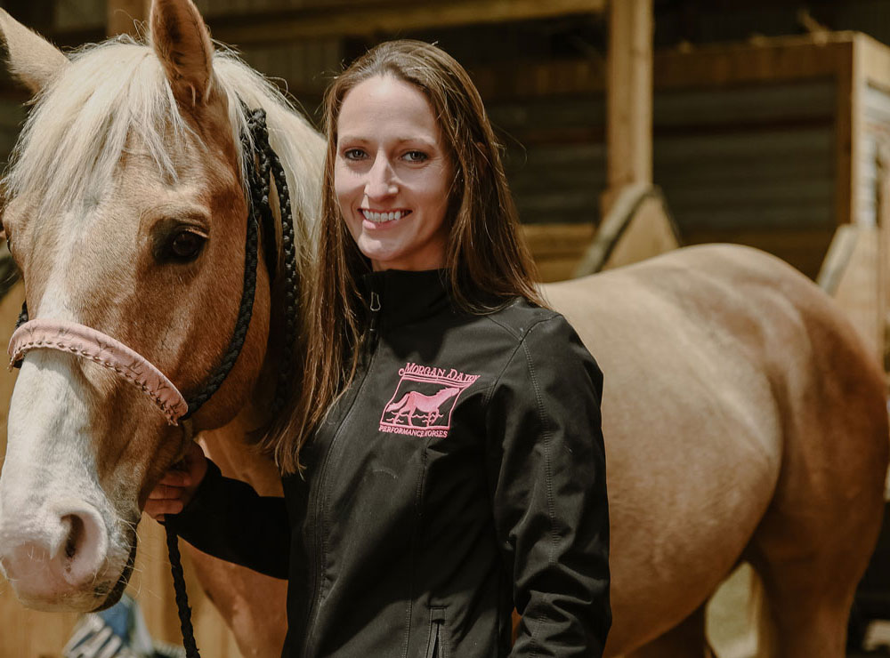 Amanda Morgan de Morgan Dairy Performance Horses vistiendo una chaqueta negra y de pie junto a su caballo rojo.